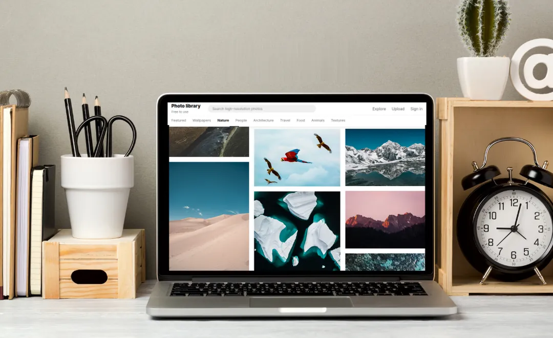 A laptop with a photo library on a desk beside a clock.